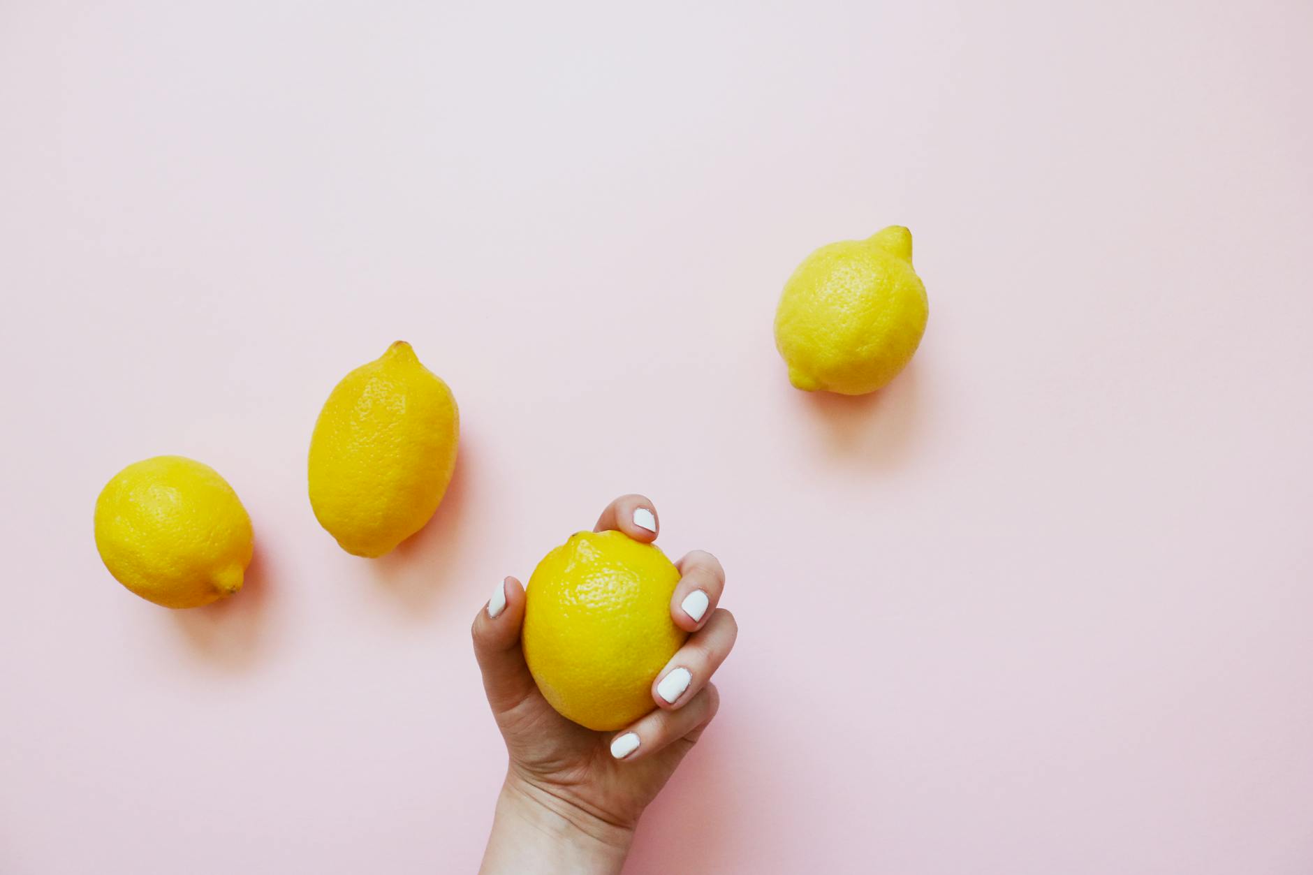 Mão com unhas brancas segurando um limão em fundo rosa suave com três limões adicionais ao redor