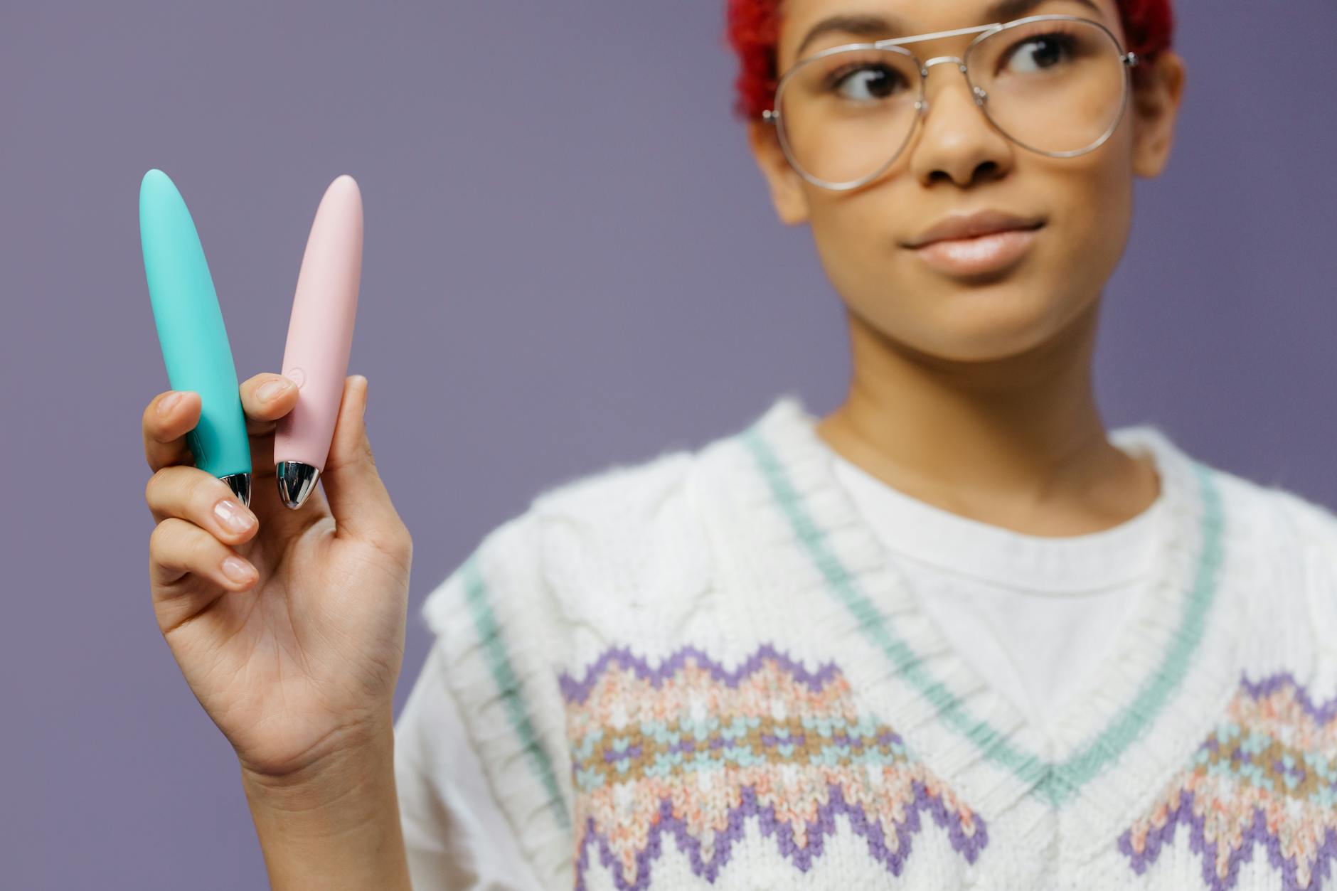 Mulher jovem com óculos segurando dois vibradores coloridos contra fundo roxo
