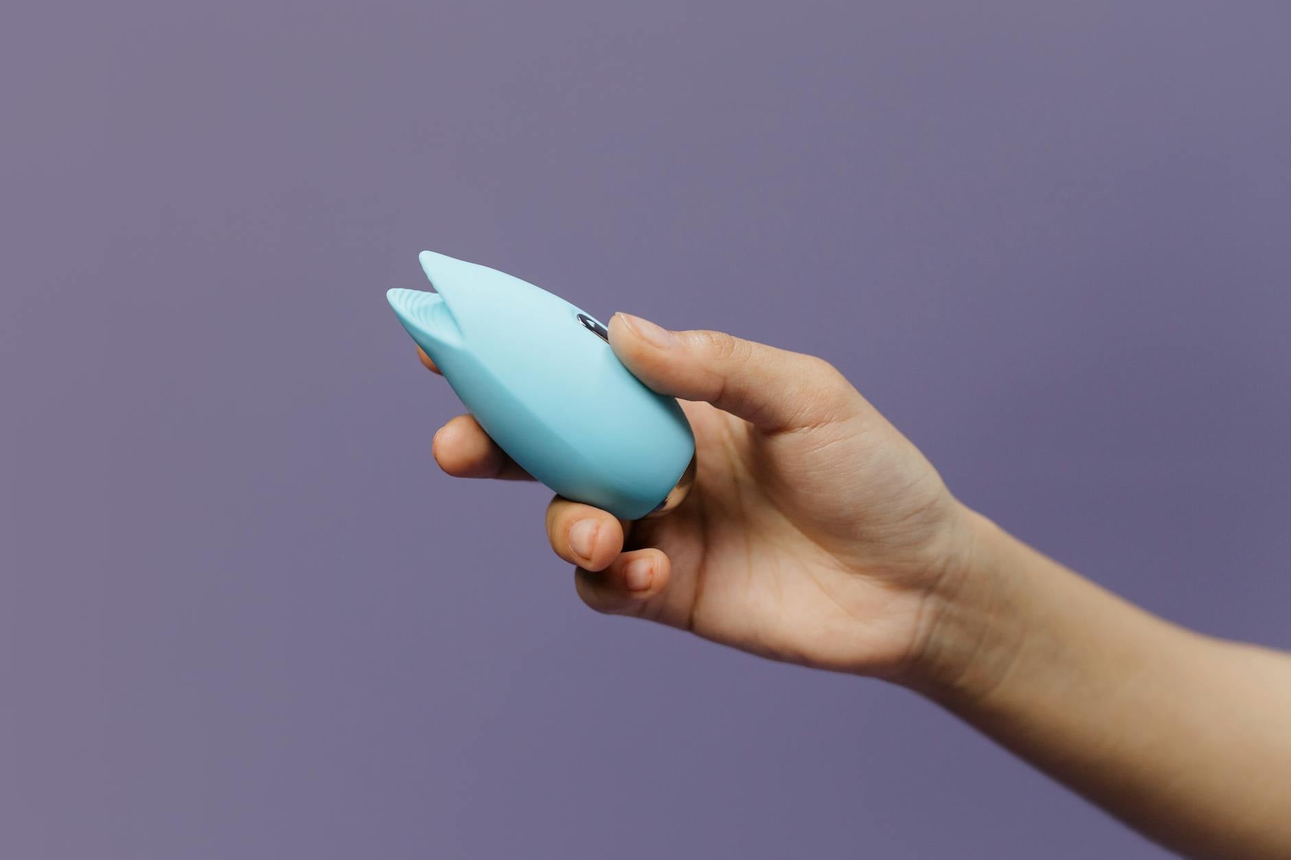 Mão segurando um vibrador de silicone azul contra fundo roxo sólido, promovendo amor próprio e sexualidade