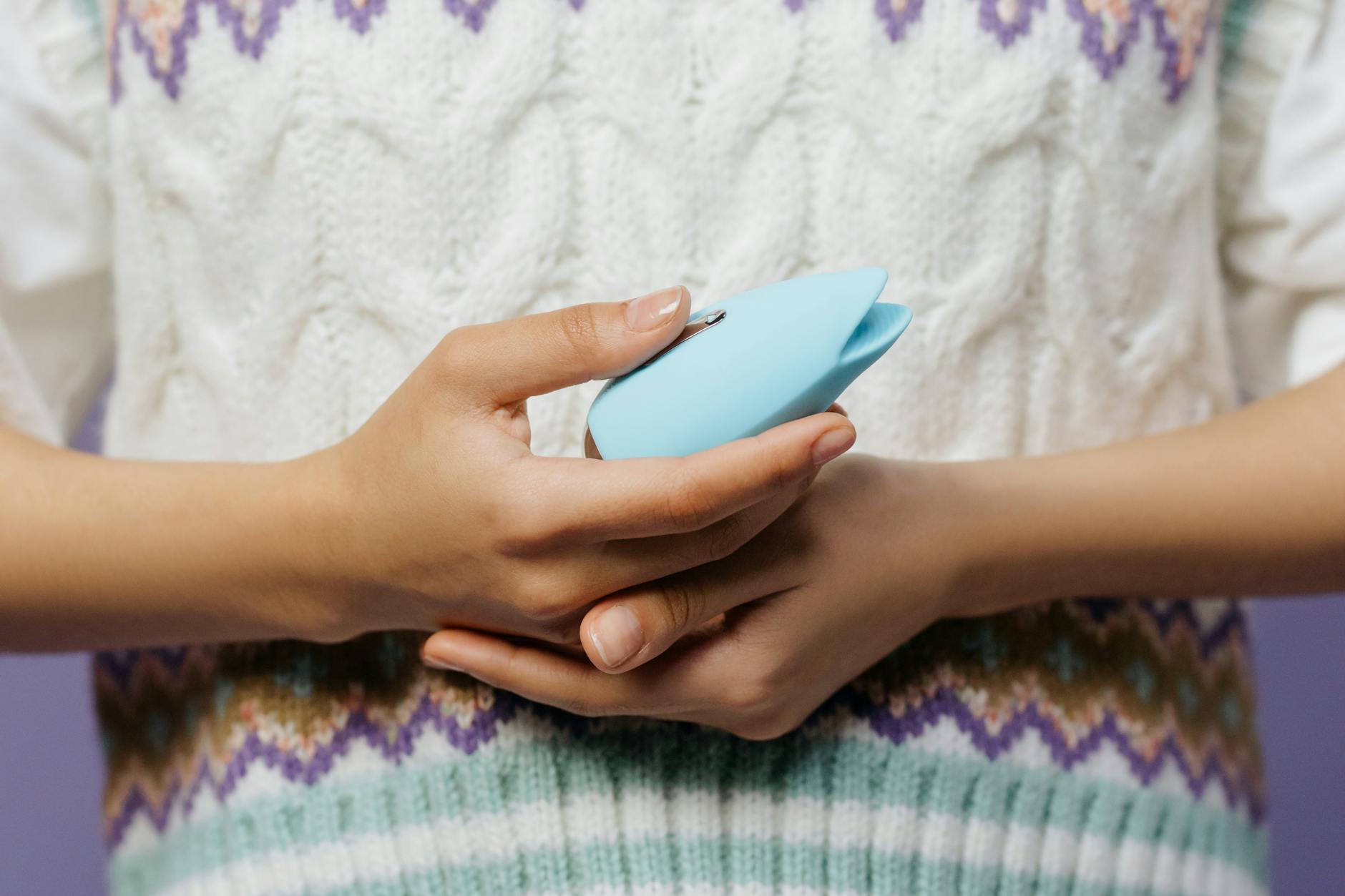 Detalhe de mãos segurando um vibrador azul contra um suéter de tricô