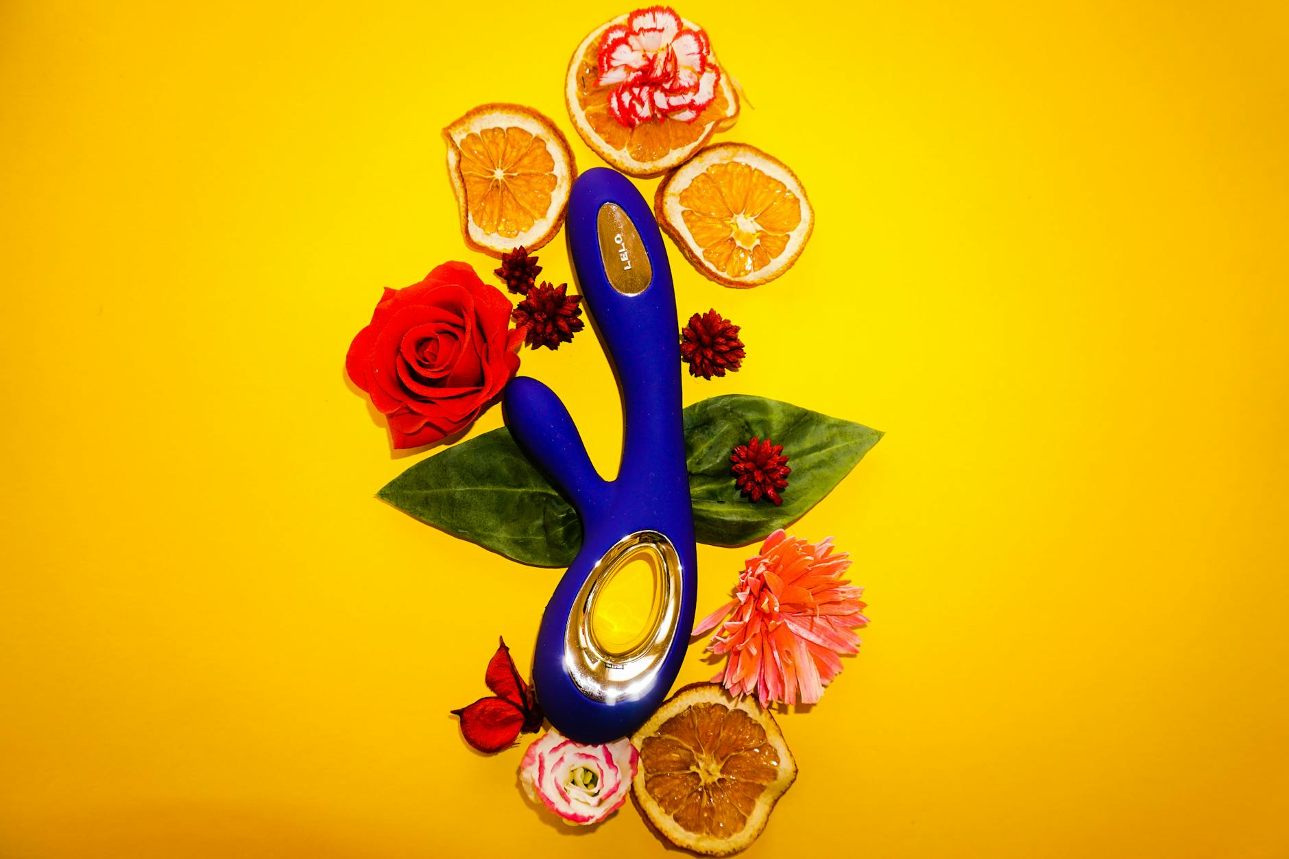Vibrador de silicone azul rodeado de flores e fatias de laranja contra fundo amarelo.